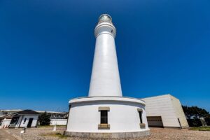 濃い青空を背景に、真下から見上げた犬吠埼灯台。白い円筒形の塔がまっすぐ伸び、頂部に黒い手すりの回廊とガラス張りの灯室がある。塔の足元は円形の白い基部で、四角い格子窓が二つ見える。左手前に白い平屋の付属舎、右手に白い箱型の建物、地面は砂利敷き