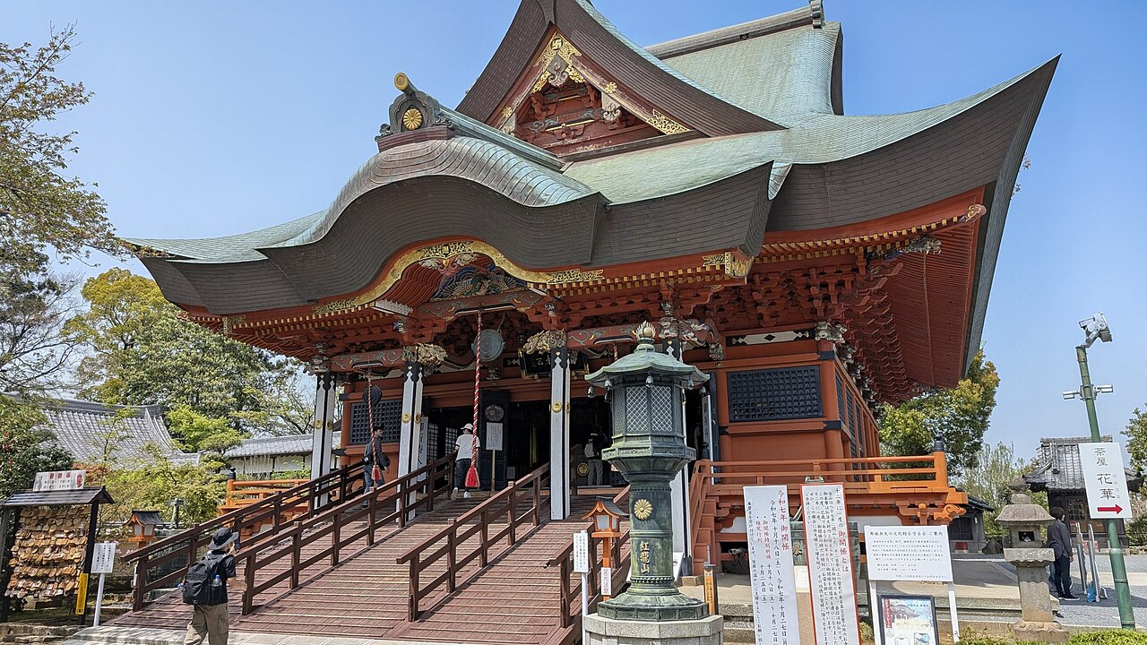 布施弁天東海寺の本堂。朱塗りの柱と反った銅板屋根を持つ建物へ、手すり付きの階段が正面からまっすぐ伸びている。階段の下には由緒書きの立て看板と絵馬掛けが並び、右手に「茶屋 花華」への矢印看板が立つ