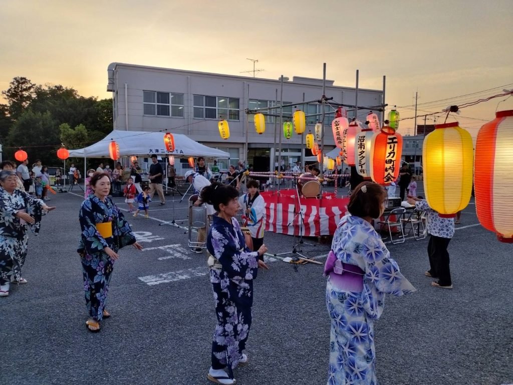 沼南庁舎の駐車場に提灯が張り巡らされ、浴衣姿の女性たちが輪になって踊っている。中央奥に紅白幕を巻いた太鼓の台があり、提灯には協賛団体名が書かれている