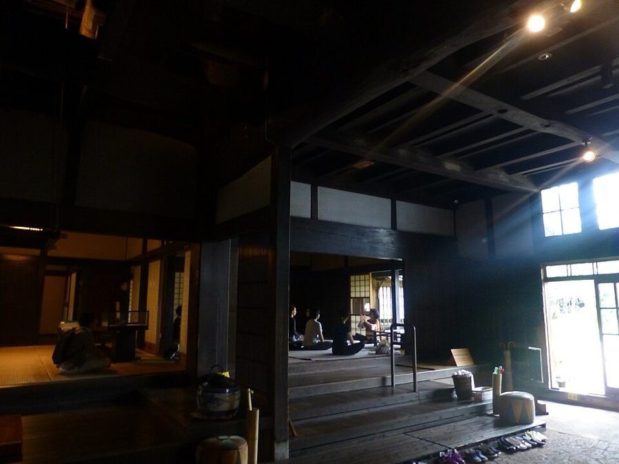 旧吉田家住宅の主屋内部。太い黒い梁と柱が組まれた高い吹き抜けの土間から、一段上がった畳の座敷を見通した屋内風景