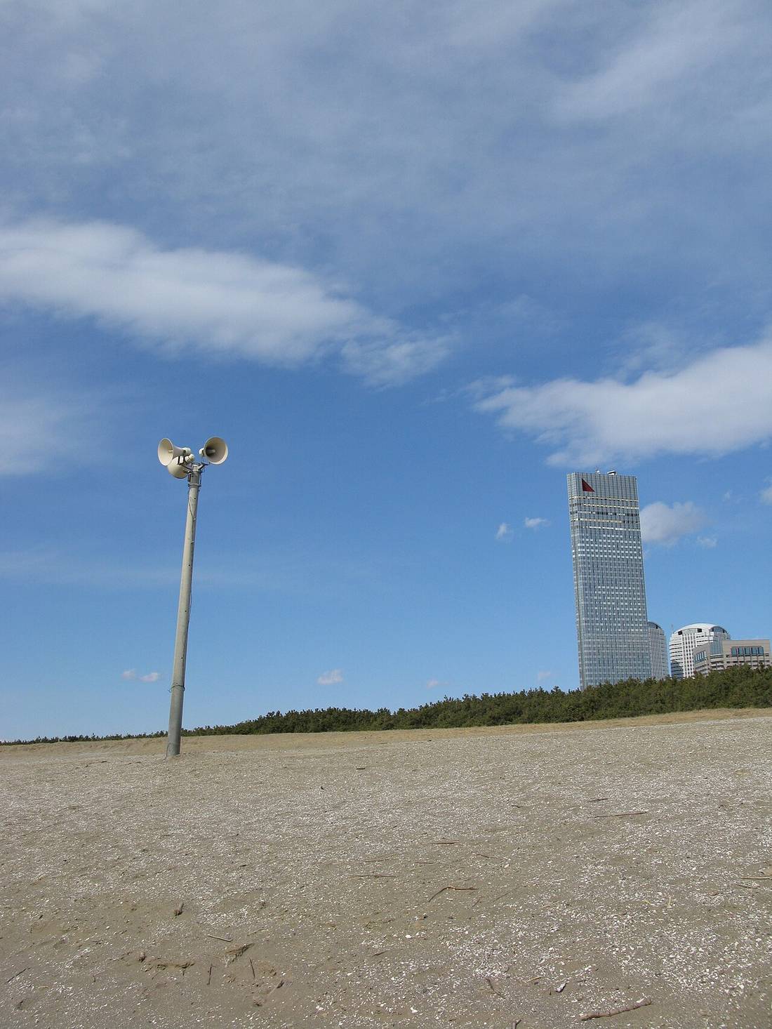 幕張の浜の砂浜に立つ拡声器付きの支柱。右奥に高層ビルとホテル、手前は貝殻混じりの砂地