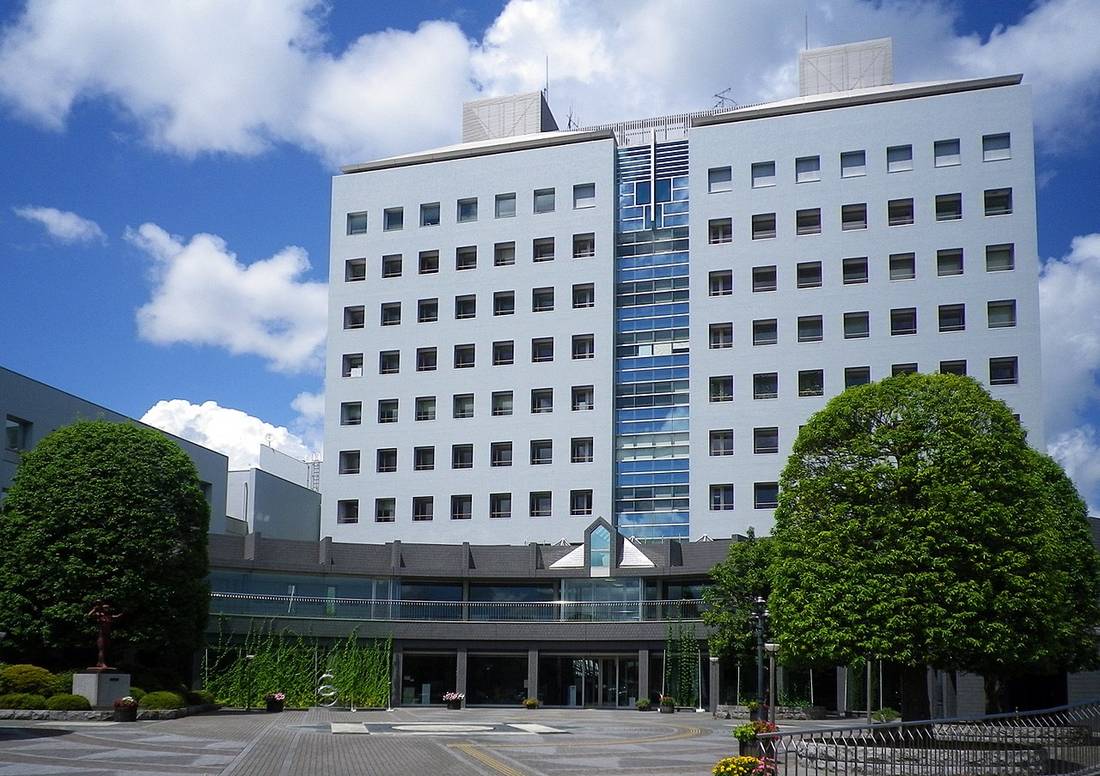 茂原市役所の庁舎。白い外壁に規則正しく窓が並ぶ高層棟、中央にガラス張りの吹き抜け、手前に丸く刈り込まれた街路樹とタイル敷きの広場