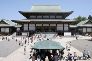 成田山新勝寺の大本堂。緑青色の屋根をもつ大きな堂の前に石段が広がり、手前の屋根付きの常香炉に人が集まっている