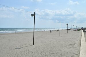 白里海岸の広い砂浜。左手に打ち寄せる波、砂浜に拡声器を載せた細い支柱が等間隔に立ち、右奥に人影と海の家