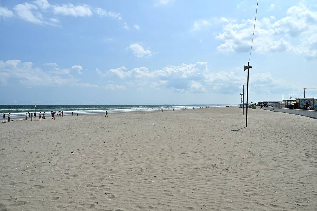 白里海岸の砂浜を海側から見た風景。遠くの波打ち際に十数人が並んで歩き、右奥に海の家らしい建物と電柱が並ぶ