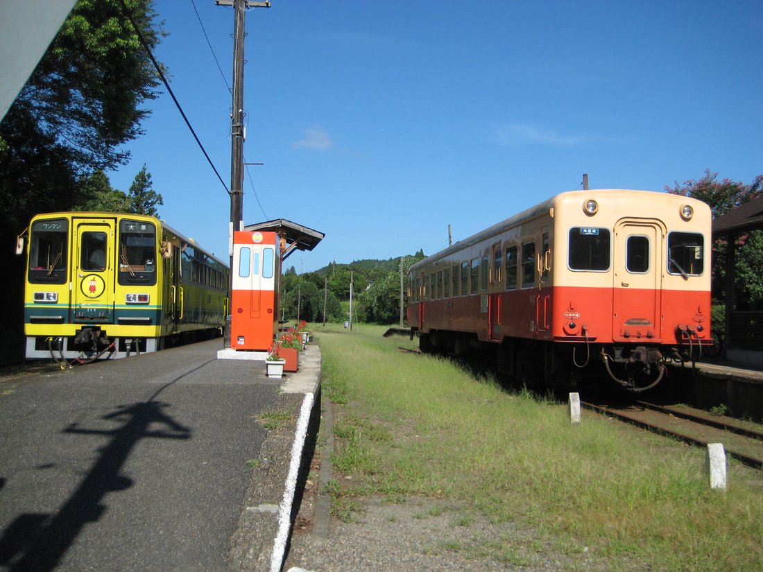 上総中野駅のホームで向かい合う2両の気動車。左は黄色いいすみ鉄道の車両で方向幕に「大原」「ワンマン」、右はクリーム色と朱色の小湊鐵道の車両。中央にオレンジ色の小屋が立ち、線路は草に覆われている
