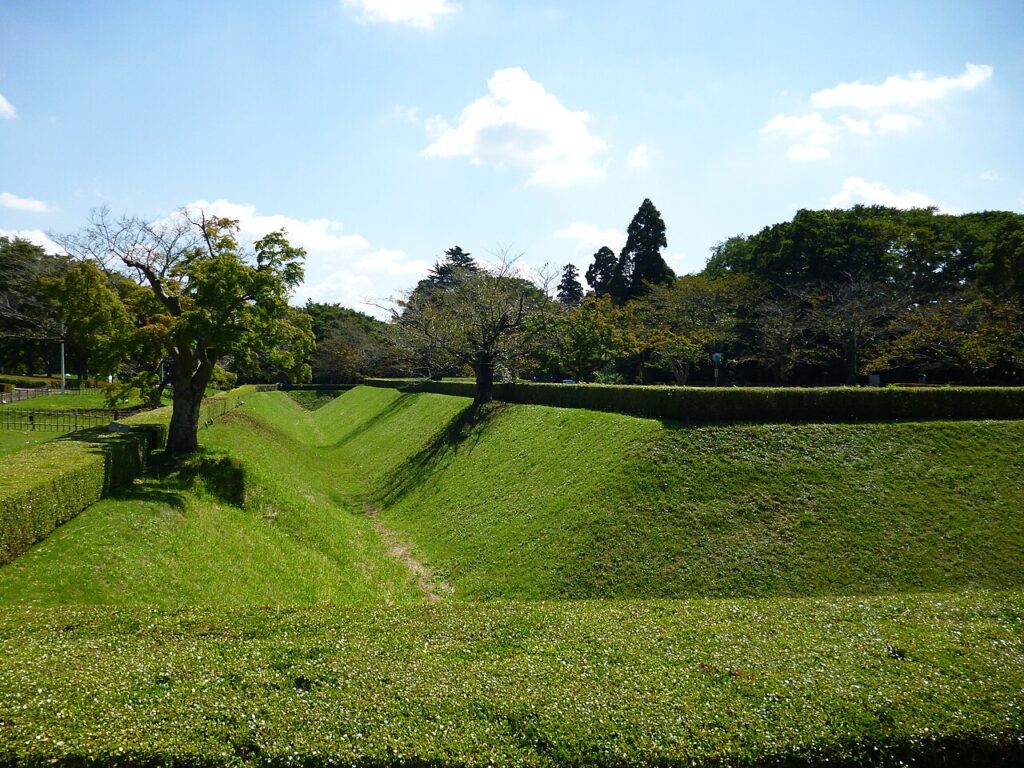 芝に覆われた城跡の空堀と土塁。手前から奥へV字に切れ込んだ堀が伸び、両側の土塁の上には刈り込まれた生垣と桜とみられる落葉樹が並ぶ。青空に雲が浮かぶ