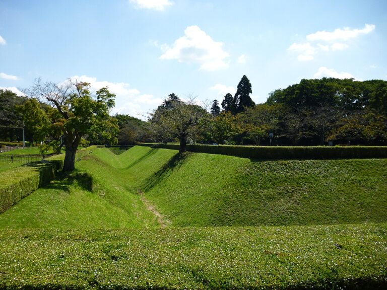 芝に覆われた城跡の空堀と土塁。手前から奥へV字に切れ込んだ堀が伸び、両側の土塁の上には刈り込まれた生垣と桜とみられる落葉樹が並ぶ。青空に雲が浮かぶ