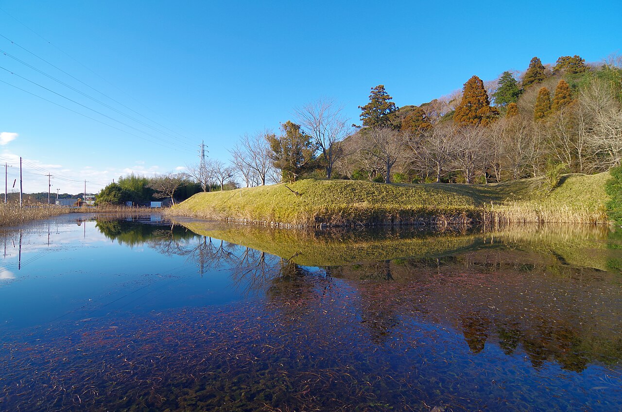 冬の水堀。手前に水面が広がり、対岸に草に覆われた土塁がゆるやかな稜線を描く。奥は落葉した木立と常緑樹の斜面、左手には送電線と鉄塔が見える