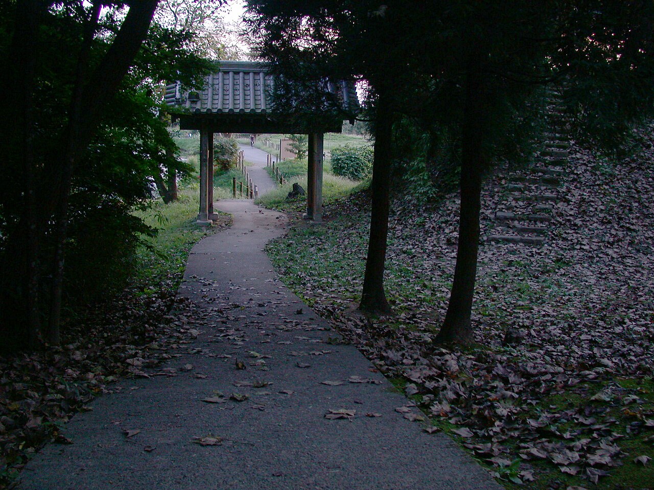 木立に囲まれた薬医門。瓦屋根を2本の柱が支え、門の下を舗装された細い園路が通り抜けている。右手の土塁の斜面には落ち葉に埋もれた階段が上へ続く