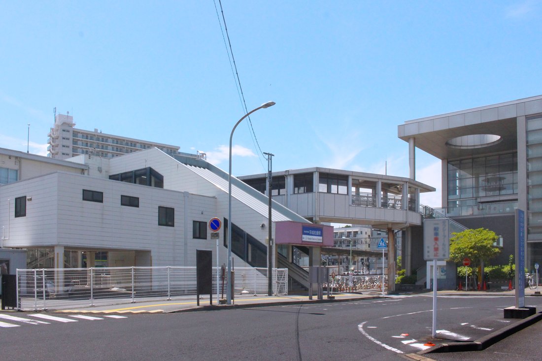 京成佐倉駅北口の駅前。屋根付きの跨線橋と長い階段、右手にガラス張りの建物、奥に駐輪場、手前は横断歩道と車道が広がる快晴の風景