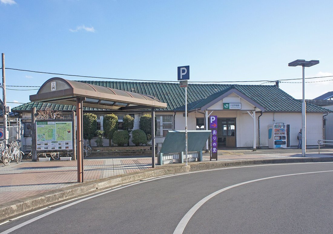 JR総武本線・八日市場駅の駅舎を駅前ロータリーから見た写真。緑色の瓦屋根の低い平屋で、左手にタクシー乗り場のアーチ状の上屋、右手に自動販売機があり、駅前に「祈りの里」と書かれた紫の看板が立っている