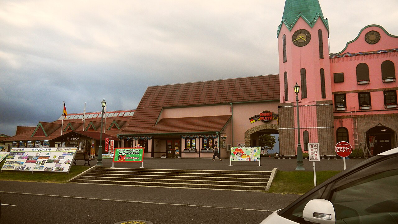 東京ドイツ村のマルクトプラッツ。緑の尖塔と時計を持つピンクの塔、木造の建物が並び、手前は広い舗装路になっている。右手前に乗用車が停まり、「駐車禁止」「防火水槽」の標識が立つ