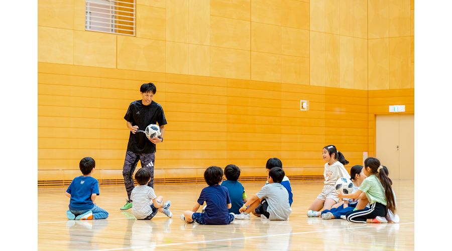 体育館で子どもたちが座り、波戸康広さんがボールを使って教える様子