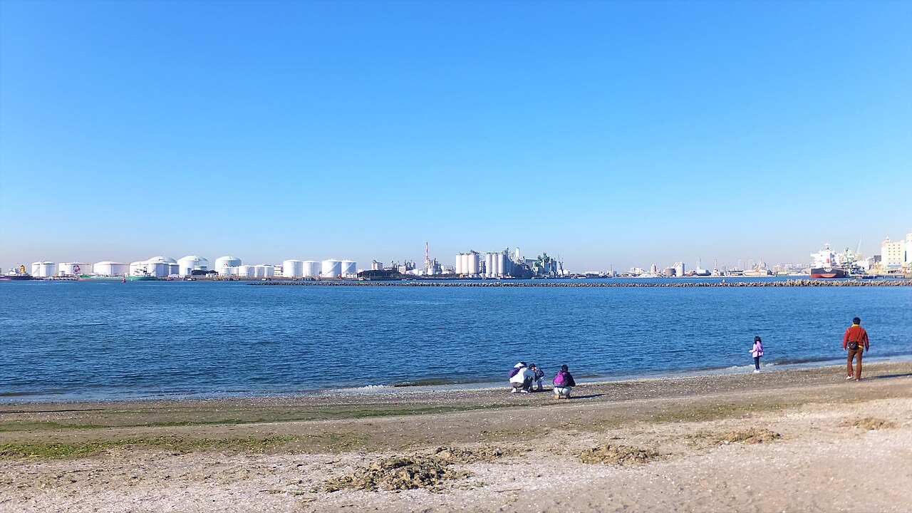 千葉ポートパークのビーチプラザ。砂浜の波打ち際に数人がしゃがんでおり、沖には消波堤、対岸には石油タンクと工場の設備が並んでいる。