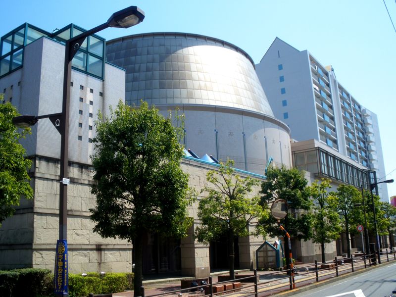 ギャラクシティの外観。円錐台形の銀色のプラネタリウムドームが建物中央にせり出し、手前の歩道には街路樹と街灯が並ぶ