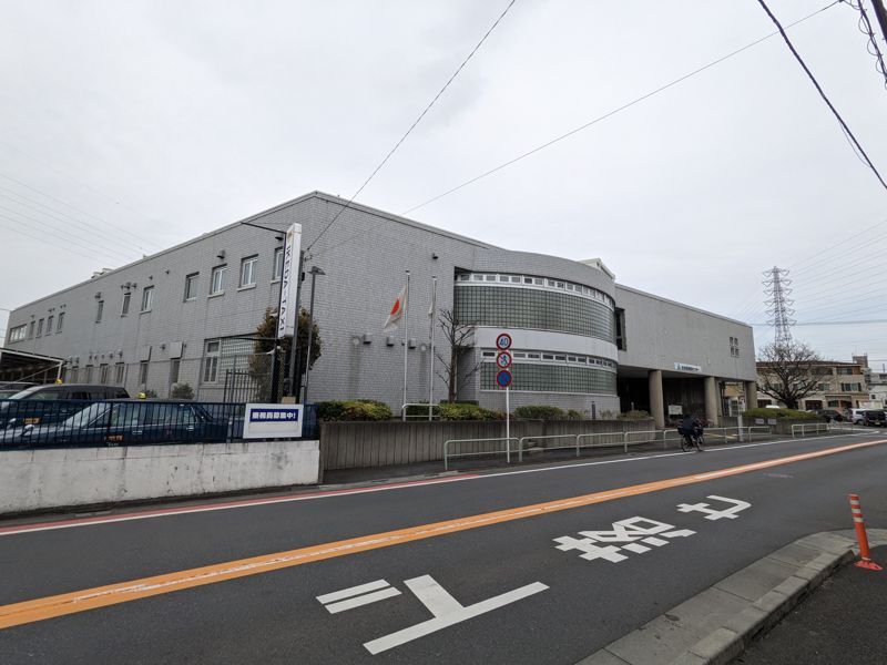 足立区保塚センターを別角度から見た外観。手前の道路に横断歩道の白線と黄色いセンターラインが走り、建物の右手前に施設名の看板が立つ