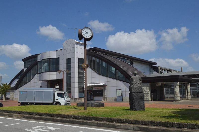 JR八街駅の駅舎。曲線の屋根が連なる2階建ての建物で、手前に時計塔と石像が立つ