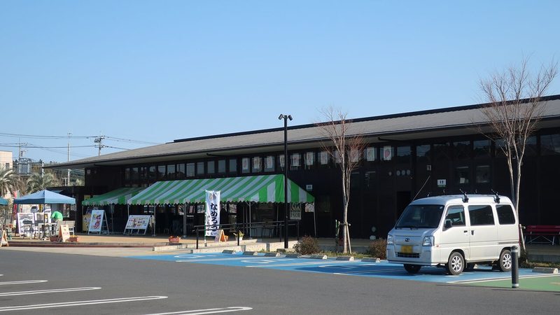 国道126号から見た道の駅みのりの郷東金。黒い横長の東金マルシェの建物の前に緑と白のテントとのぼりが立ち、手前は駐車場