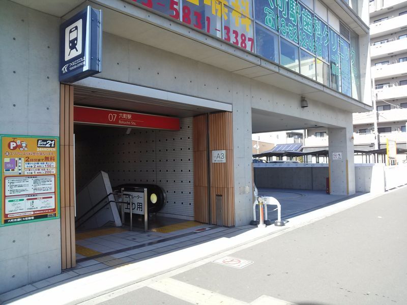 つくばエクスプレス六町駅のA3出口。ビルの1階に開いた地下への階段口で、赤い庇に「07 六町駅」の表示、右手の壁に「A3」の標識が付く