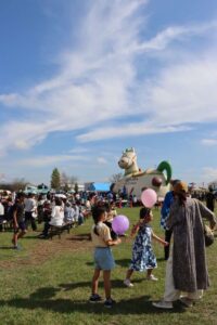 白井市ふるさとまつりの会場の白井総合公園。晴れた空の下、芝生の広場に子どもたちが風船を持って歩き、奥に馬の形のふわふわ遊具とテントの出店が並んでいる