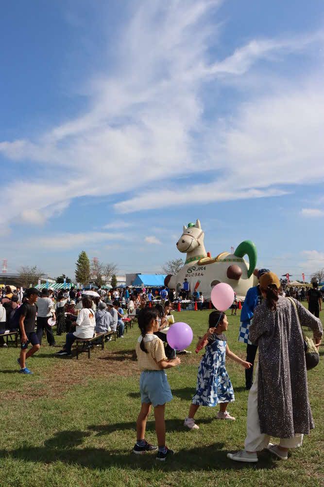 白井市ふるさとまつりの会場の白井総合公園。晴れた空の下、芝生の広場に子どもたちが風船を持って歩き、奥に馬の形のふわふわ遊具とテントの出店が並んでいる