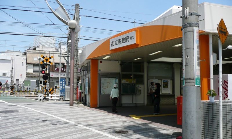 西武新宿線・都立家政駅南口。オレンジ色の庇の下に改札があり、左手に踏切、右手に赤いポストが写る