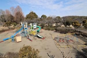 青空の下、すべり台やネットのある大型遊具と砂地の広場が写った写真