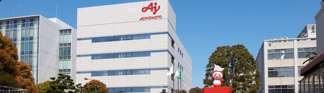 「AJINOMOTO」のロゴがある白い工場の建物と、旗を持つアジパンダの像