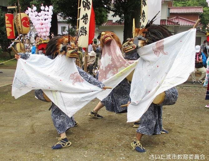白い布を垂らし腰に太鼓を付けた3頭の獅子が土の境内で舞い、後ろに花の飾りを付けた竿、幟、見物人が見える写真