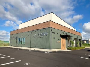 青空の下、緑の外壁に木目調の帯がある平屋の建物と、手前の駐車場
