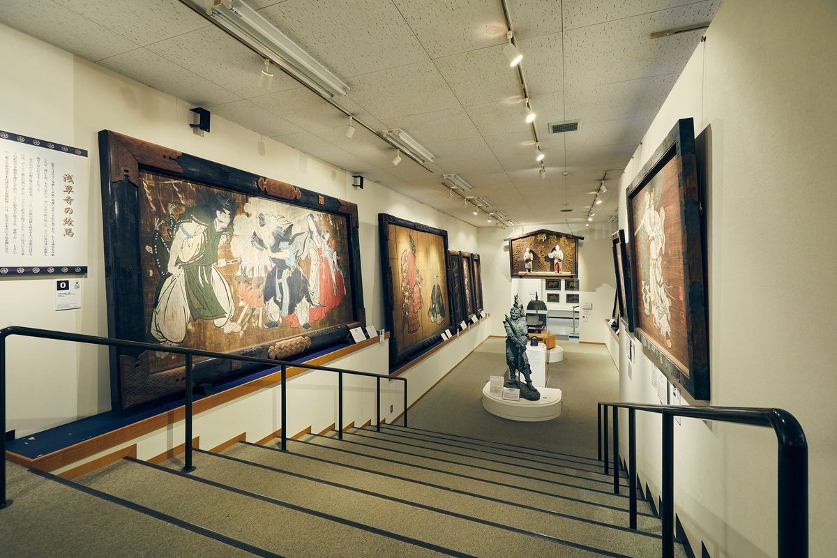 壁に大きな絵馬が並び、中央に像が置かれた絵馬堂の内部と、手すりの付いた階段