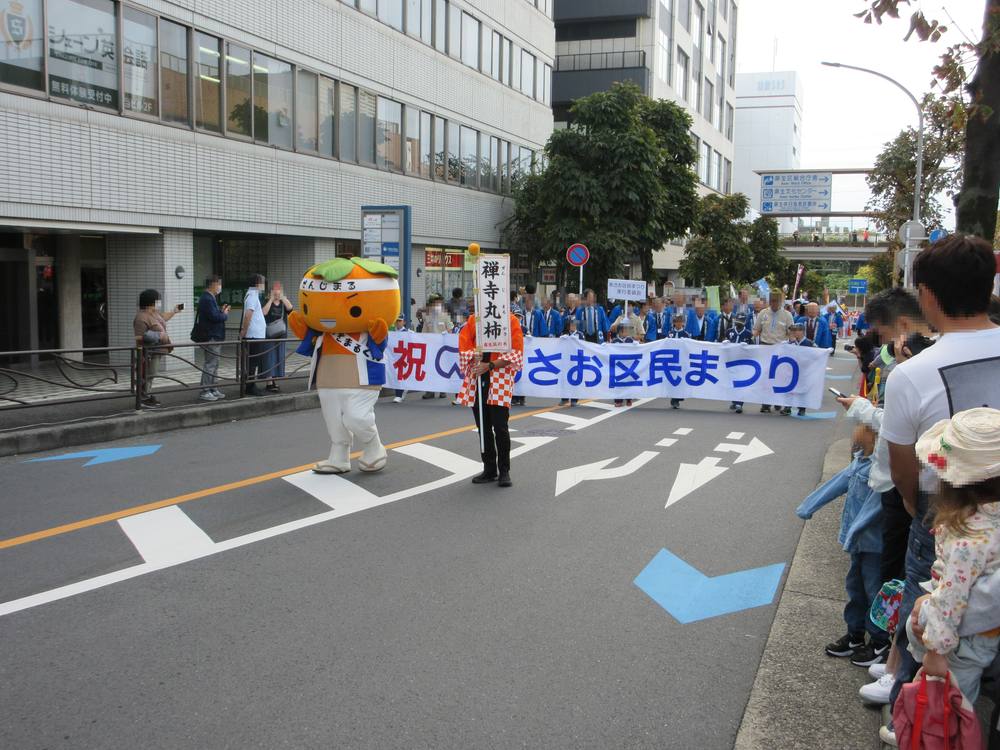2023年のあさお区民まつりのパレード。区役所前の道路を禅寺丸柿のキャラクターかきまるくんと横断幕を持った列が進む