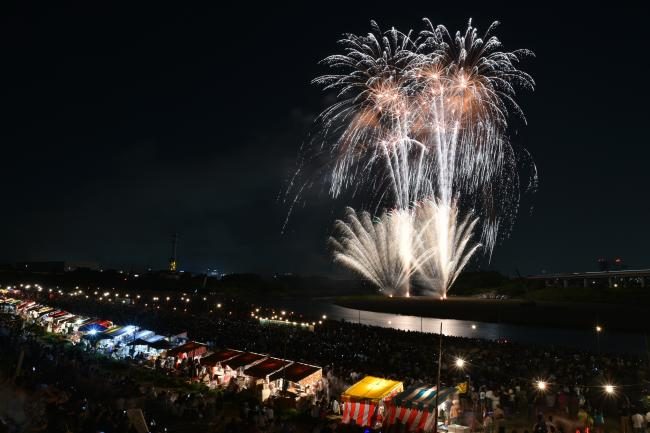 相模川の河川敷に屋台の明かりが並び、その奥で花火が上がっている夜景