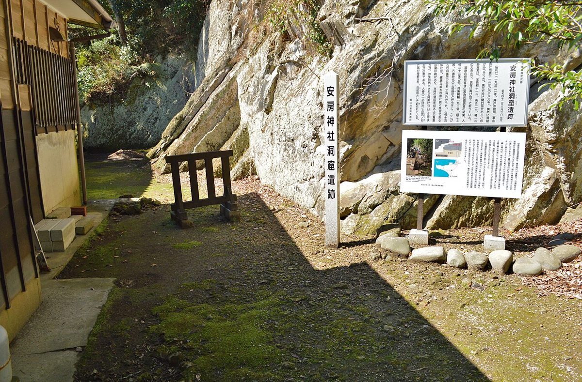 安房神社の境内にある洞窟遺跡の跡地と説明板。岩壁の前に小さな鳥居が立つ