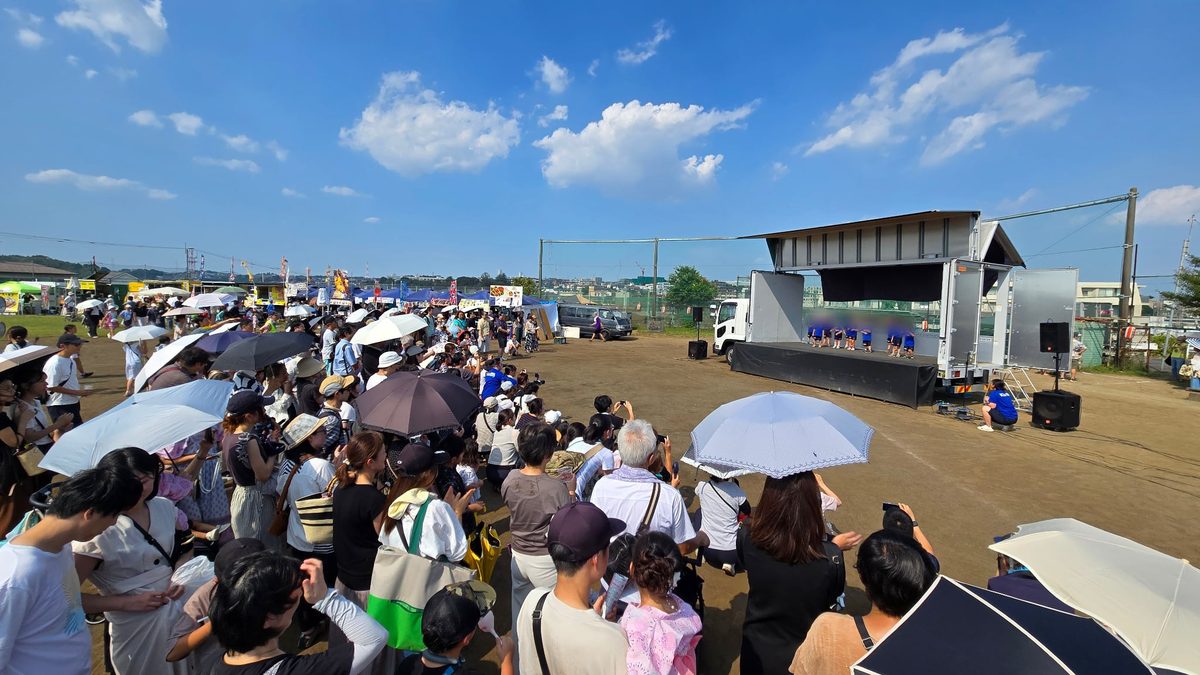 青空の下、トラックステージで演技する子どもたちと、日傘をさして見守る大勢の観客