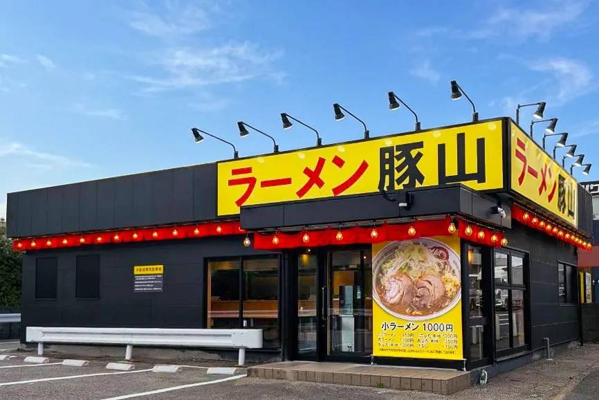 黒い平屋の店舗に黄色い「ラーメン豚山」の看板。入口の横に「小ラーメン 1000円」のポスターがあり、手前に駐車場がある