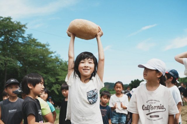 丸いチーズを両手で頭の上に持ち上げて笑う子どもと、周りの子どもたち