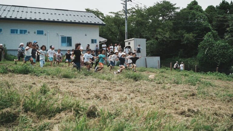 牧草地の斜面を子どもたちが一斉に駆け下りている様子。奥に白い牛舎の建物