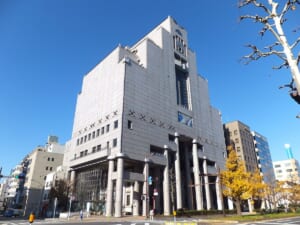 交差点の角に建つ灰色の石張りの高層の建物。下層に太い円柱が並び、手前の歩道に黄葉したイチョウがある