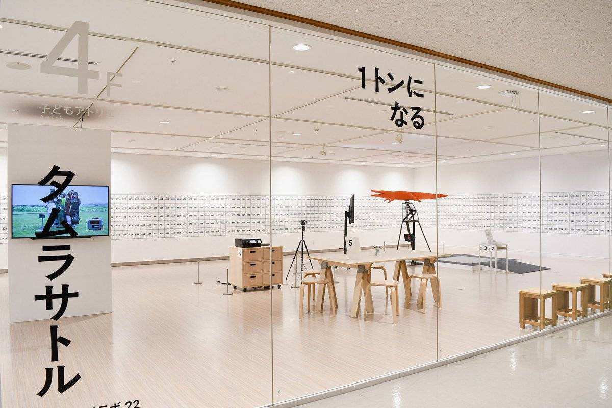 ガラス張りの4階子どもアトリエの入口。「タムラサトル」「1トンになる」の大きな文字の奥に、番号のマスが並ぶ白い壁、木の机と椅子、オレンジ色のワニの彫刻をのせた台が見える