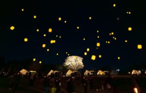 夜の芝生の上に光るランタンが多数浮かび、手前にグランピングのテントと参加者の姿が見える