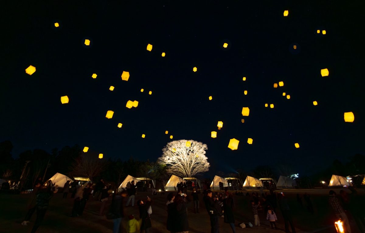 夜の芝生の上に光るランタンが多数浮かび、手前にグランピングのテントと参加者の姿が見える
