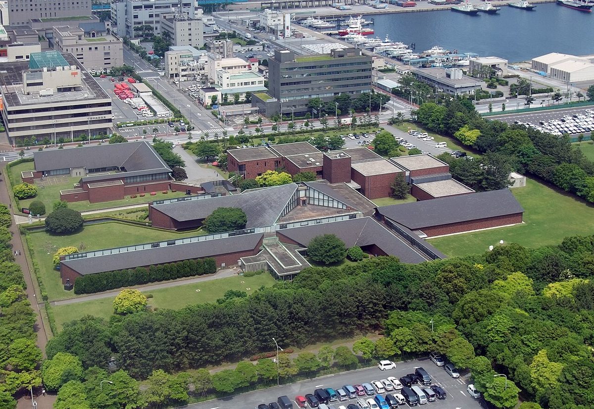 緑の芝生と木立に囲まれた、赤れんが色の壁と灰色の屋根の低い建物群を上空から写した写真。奥に港とヨットが見える