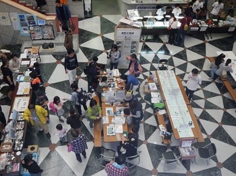 白黒の床の吹き抜けホールに長机が並び、標本や資料を囲んで子どもと大人が集まっている様子を上から写した写真