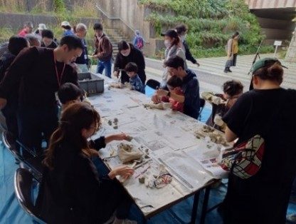 屋外の長机で親子が石を囲み、化石を掘り出す作業をしている写真