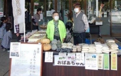 かぼちゃや袋詰めの焼き菓子を並べた販売台と、その奥に立つ出展者の写真