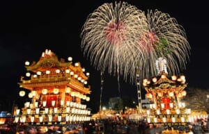 夜の御旅所で提灯に照らされた2基の屋台が並び、背後の夜空に大きな花火が開いている秩父夜祭の写真