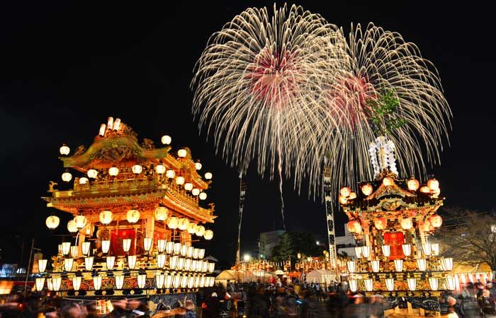 夜の御旅所で提灯に照らされた2基の屋台が並び、背後の夜空に大きな花火が開いている秩父夜祭の写真