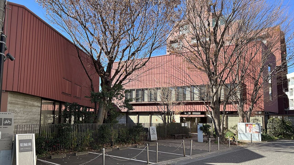 えんじ色の金属の外壁の建物が並び、葉の落ちた木の奥に入口と案内看板、手前に駐輪場が見える冬のちひろ美術館・東京